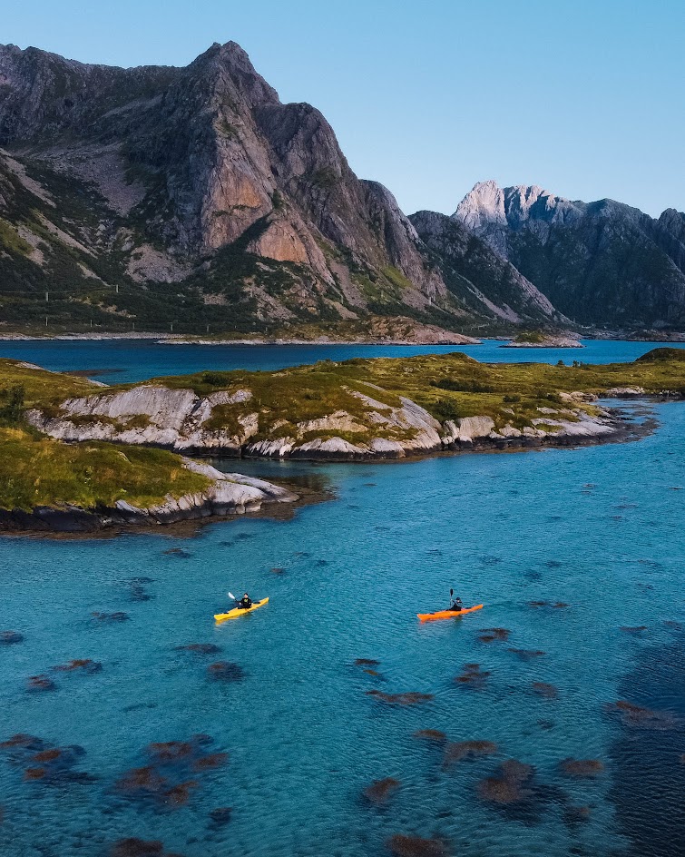 To personer i farverige kajakker padler gennem klart vand med dramatiske fjelde i baggrunden i Lofoten, Norge.